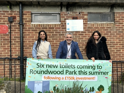 Cllr Butt and CM at Roundwood Park toilet in front of a sign saying "Six new toilets coming to  Roundwood Park this summer following a £150k investment"