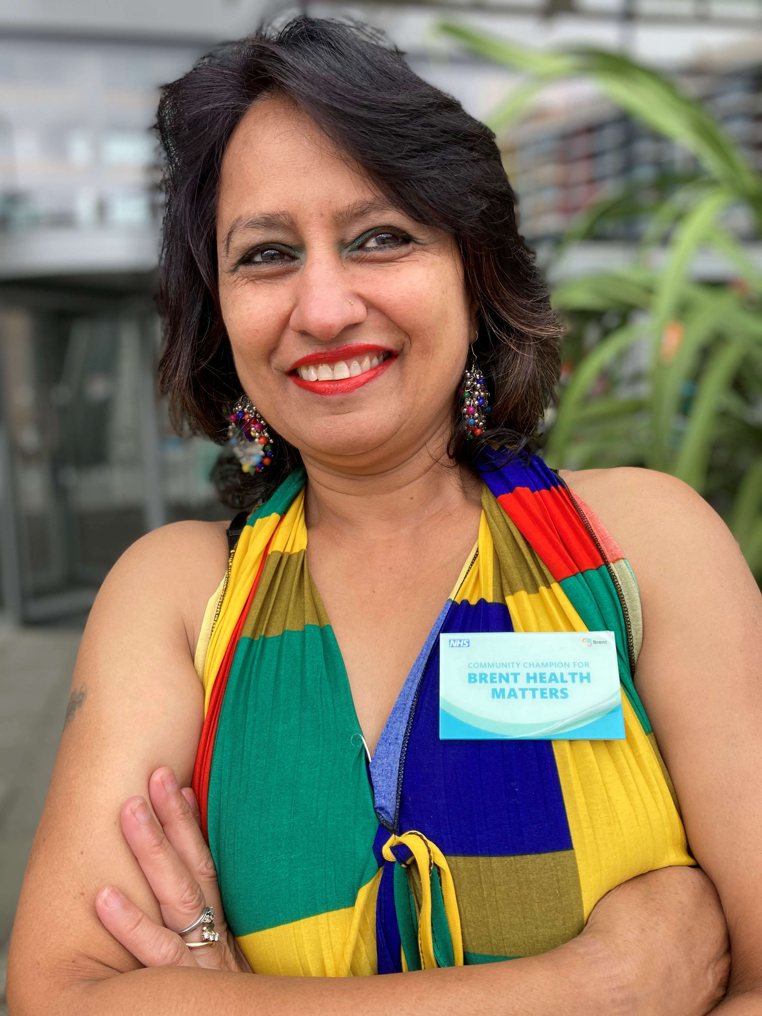 Image of a lady wearing a colourful shirt with her arms folded, wearing a Brent Health Matters badge
